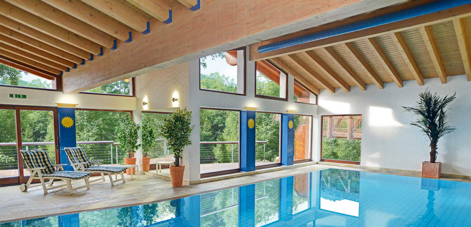 Innenpool mit großen Fensterflächen, Holzdecke mit Trägern, hellblaue Wasseroberfläche und Sitzmöbel im Hintergrund.
