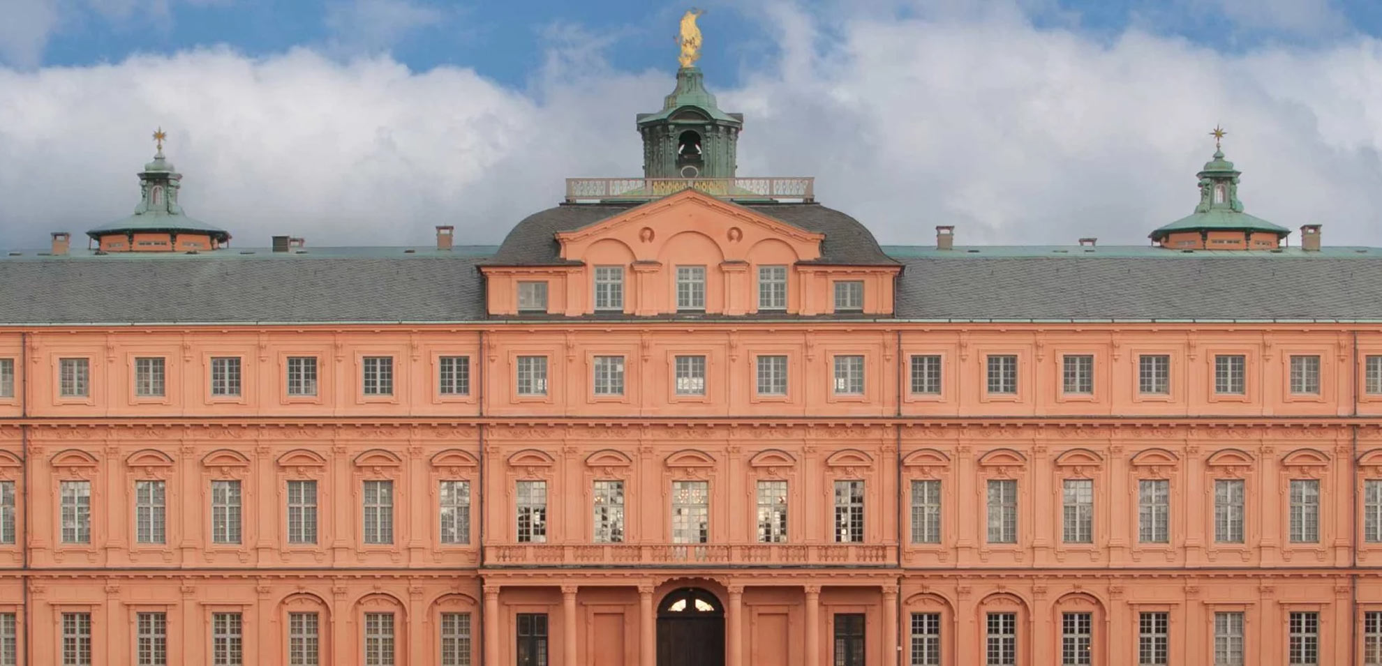 Frontale Ansicht des barocken Rathauses Rastatt mit rosafarbener Fassade, symmetrisch angeordneten Fenstern und Uhrturm in der Mitte.