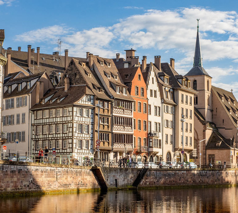 Blick auf Fachwerkhäuser am Flussufer in Straßburg, spiegelndes Wasser im Vordergrund.