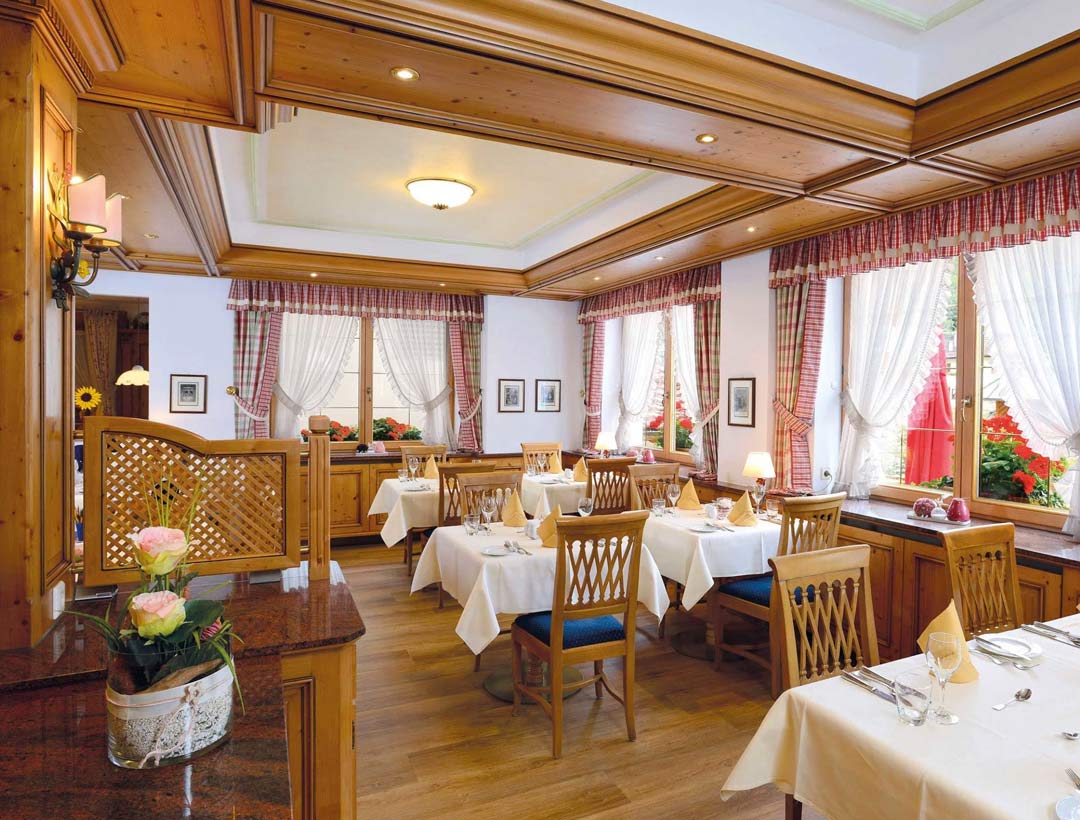 Restaurant mit roten gepolsterten Holzstühlen, Holztäfelung und weiß gedeckten Tischen, große Fenster mit Blick ins Grüne.