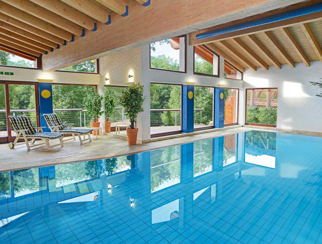 Innenpool mit Tageslicht, Holzdecke mit sichtbaren Balken, blauer Fliesenboden und Entspannungsbereich mit Liegen am Beckenrand.