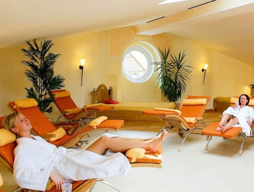 Ruhebereich mit Liegen, runder Fensteröffnung und oranger Wandgestaltung, Blick auf entspannende Atmosphäre mit Zimmerpflanzen.