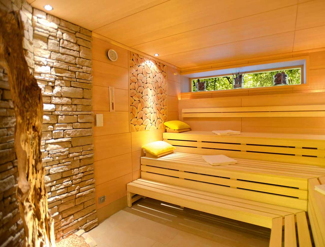 Sauna mit beleuchteter Natursteinwand, stimmungsvoller LED-Beleuchtung, hellen Holzflächen und ergonomisch geformten Bänken.