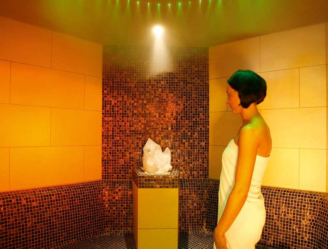 Spa-Eingang mit dekorativer Wasserstelle in einer dunklen Wandnische, farbige Lichtstimmung und klar strukturierter Raumaufteilung.