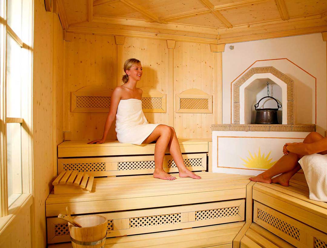 Frau in traditioneller Holzsauna auf der oberen Liegefläche, umgeben von goldgelb gefliester Wandgestaltung.