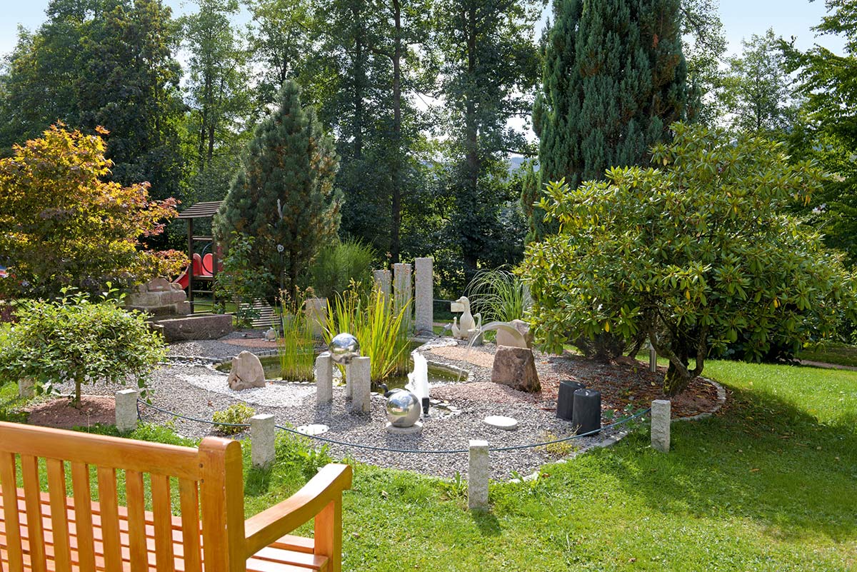 Gartenbereich mit Natursteinplatten, kleiner Holzbank, bodennaher Bepflanzung und grüner Wiese.