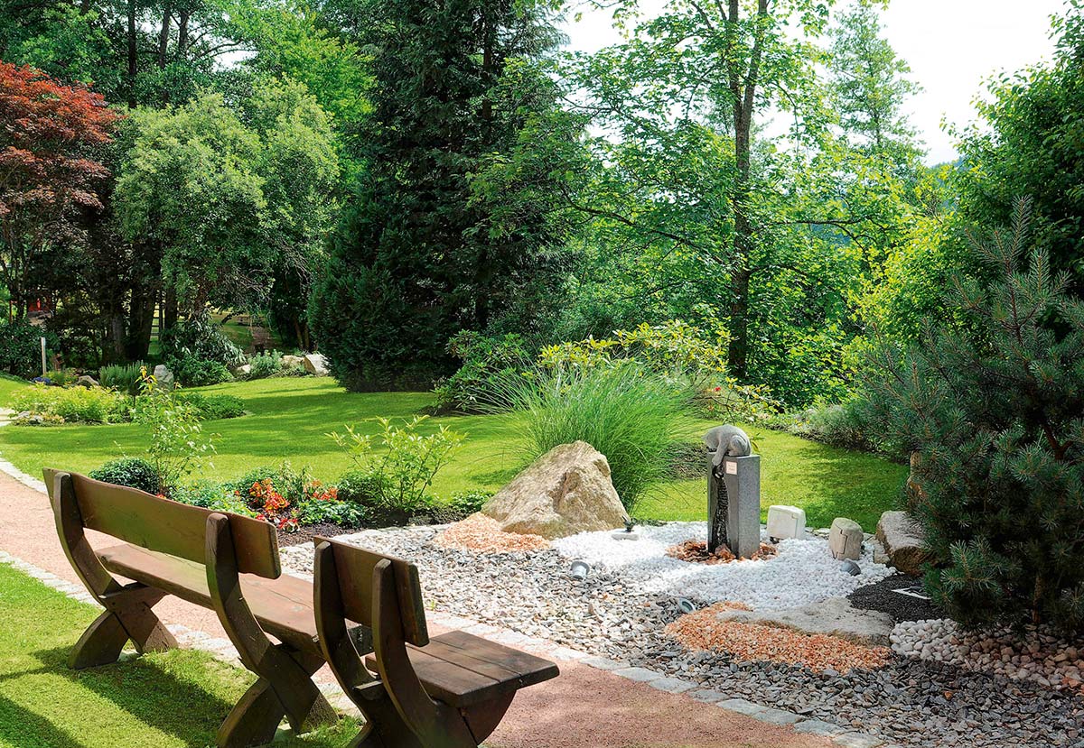 Gartenbereich mit Natursteinplatten, kleiner Holzbank, bodennaher Bepflanzung und grüner Wiese.