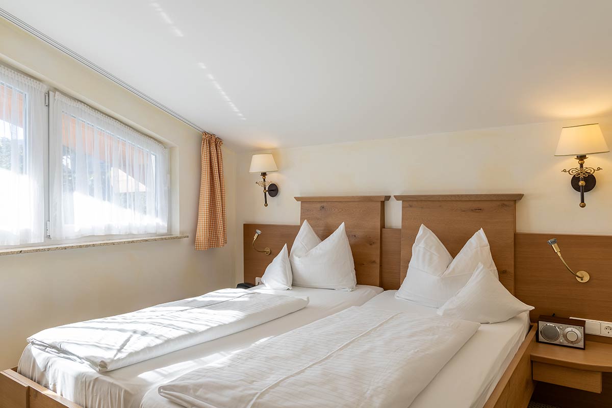Hotelzimmer mit Doppelbett, zwei Kopfkissen, Nachttischlampen und großem Fenster mit Tageslicht von rechts.