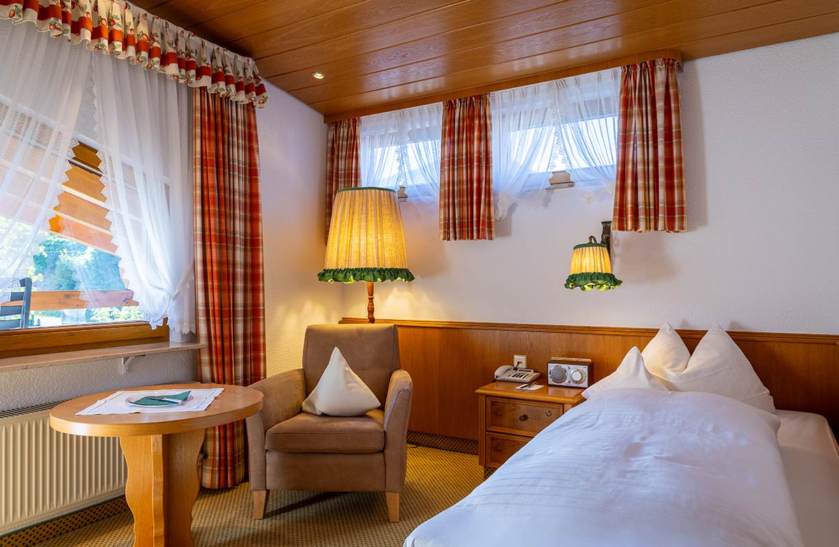 Einzelzimmer mit karierten Vorhängen, Sessel, Bett und Tisch. Rustikal-gemütlich.