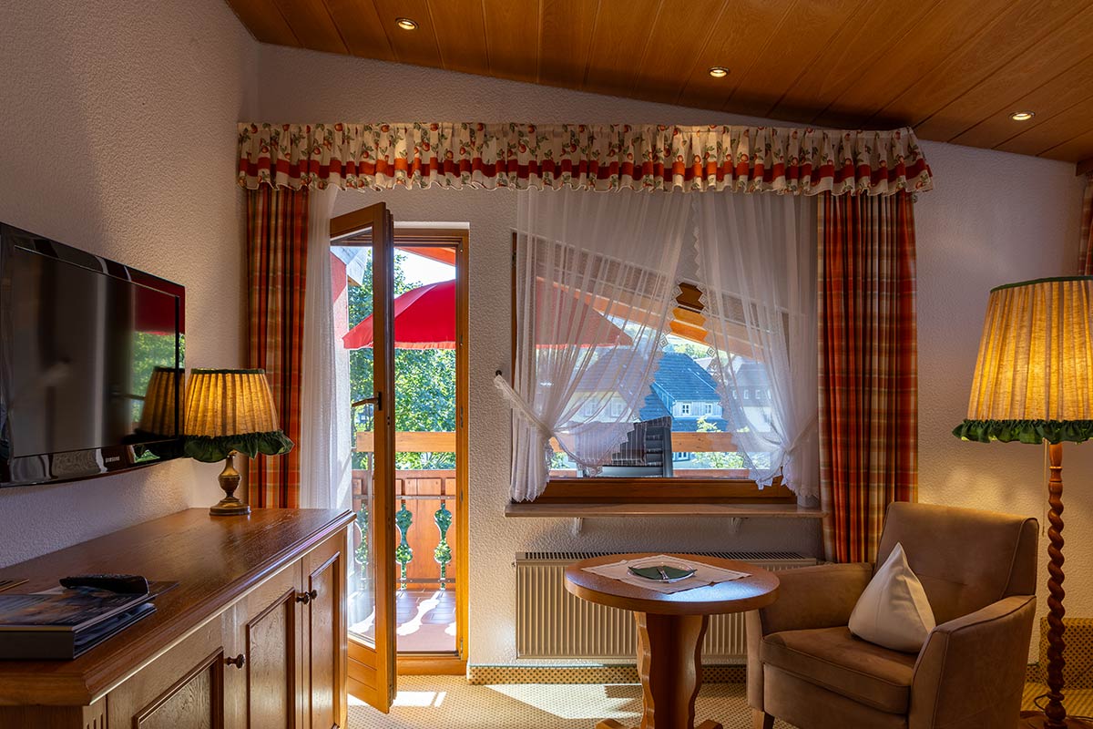 Einzelzimmer: Kleiner Esstisch mit zwei Stühlen in einer Nische, Fenster mit Blick ins Grüne, rote Vorhänge seitlich geöffnet.