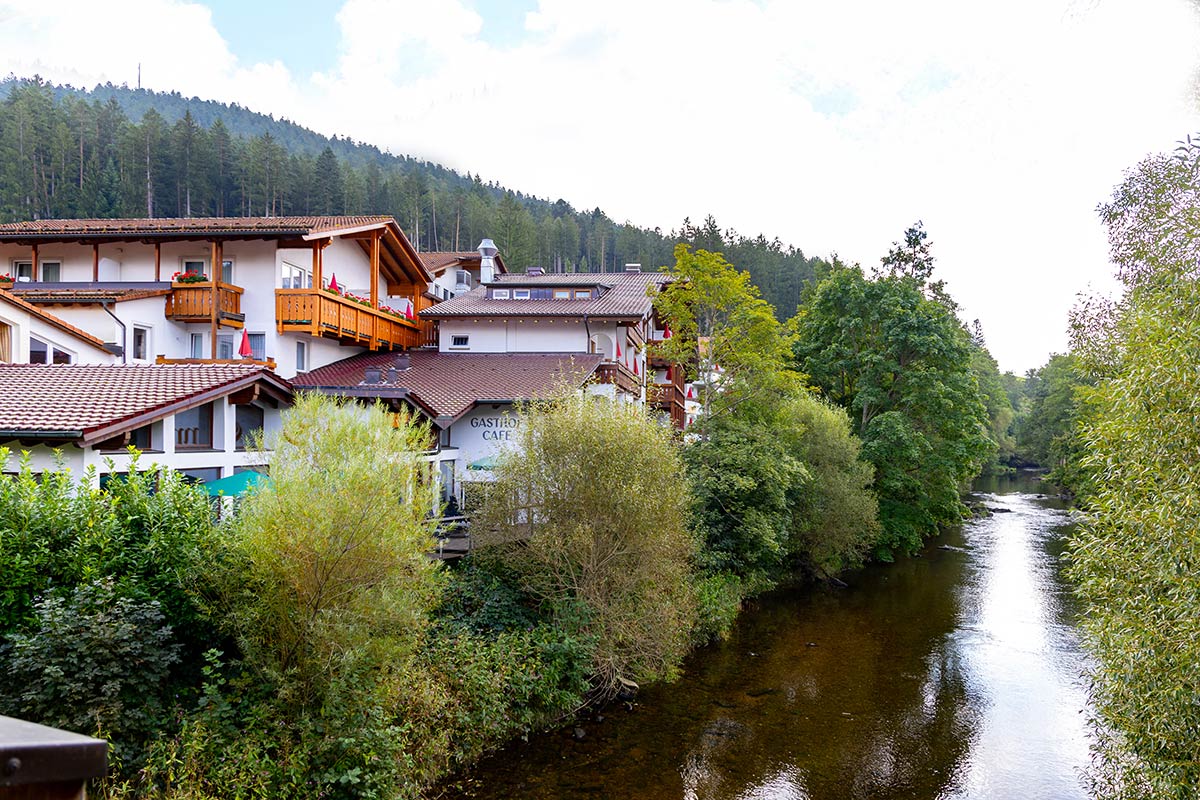 Hotelgebäude am Fluss, eingebettet in grüne Bäume, Blick über ruhiges Wasser bei Tageslicht.