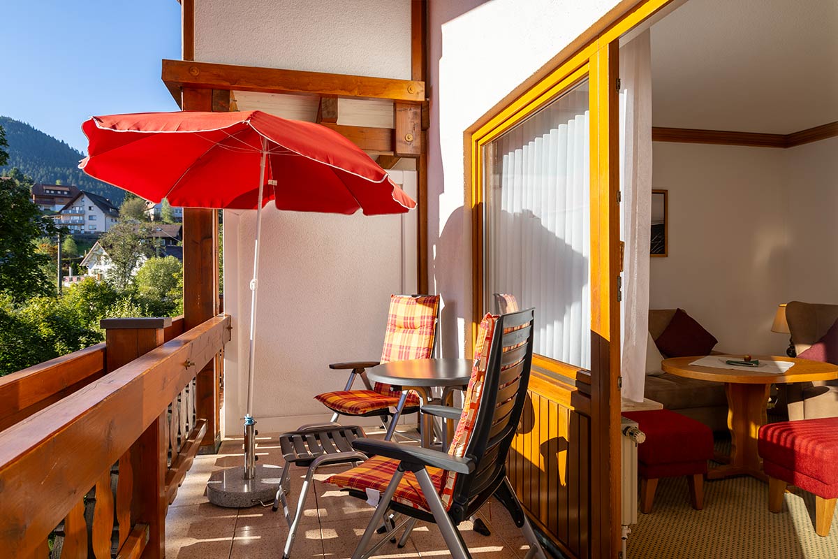 Balkonmöblierung mit zwei Stühlen, Tisch und rotem Sonnenschirm, angrenzend an Terrassentür des Zimmers.