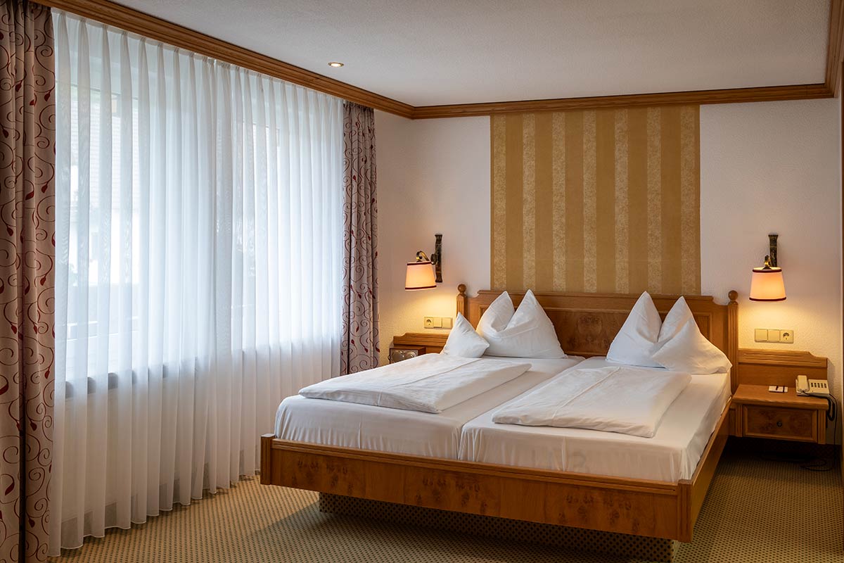 Helles Schlafzimmer mit Doppelbett, Nachttischlampen, zwei Fenstern mit weißen Vorhängen.