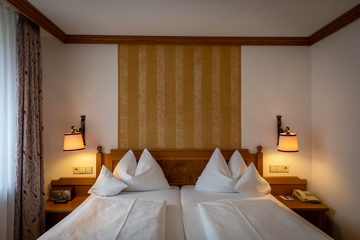 Doppelbett mit vier Kissen, Holzkopfteil, Nachttische mit Lampen, warmes Licht von rechts.
