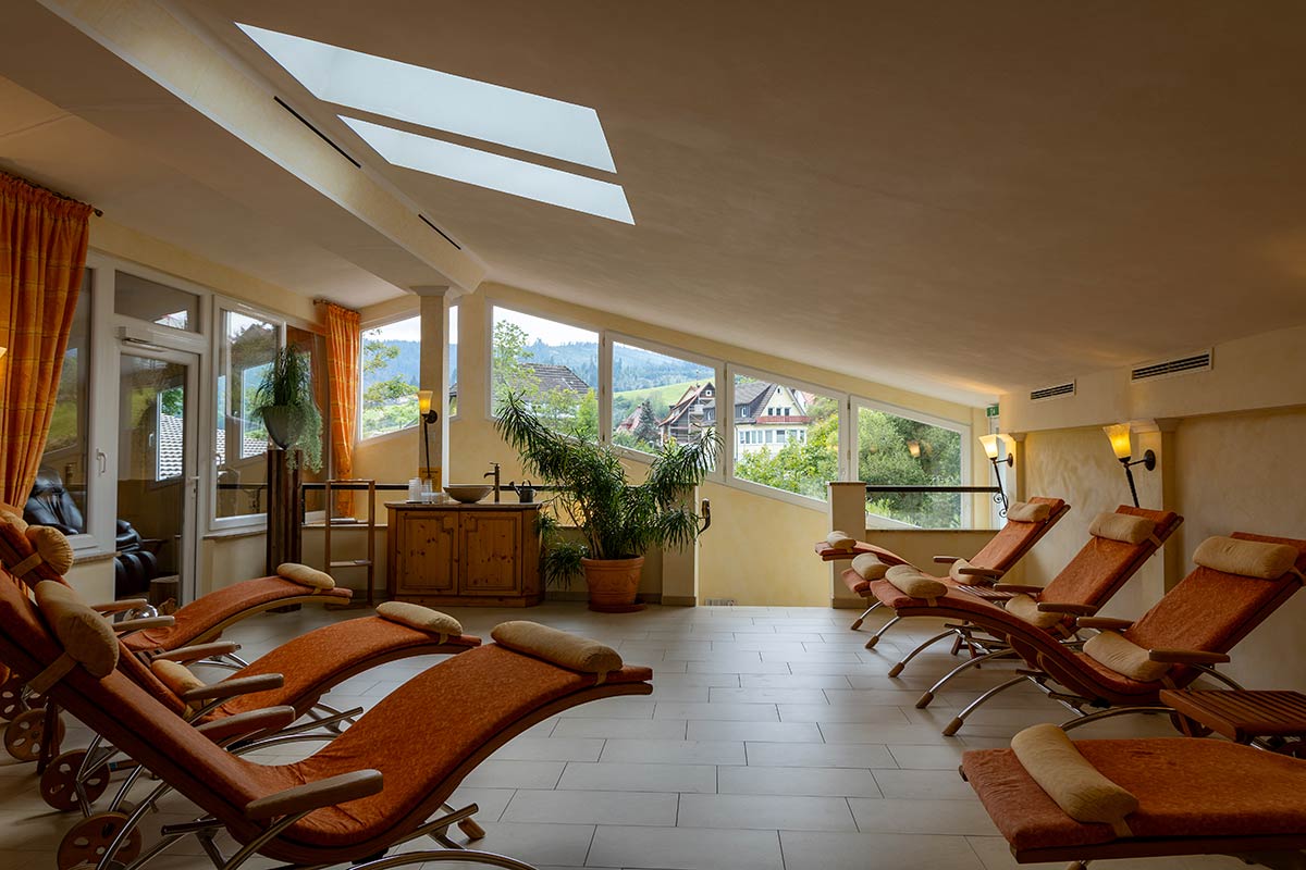 Ruhebereich mit mehreren Liegestühlen aus Holz, großen Fenstern und Blick auf Garten oder Außenanlage.