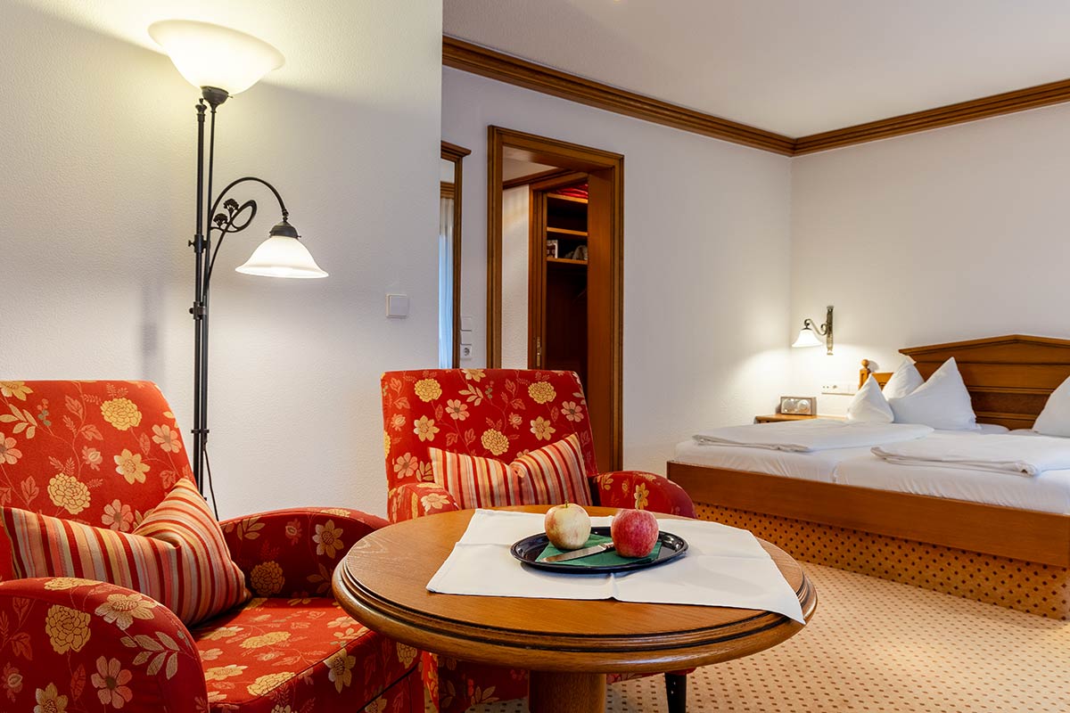 Doppelzimmer mit Doppelbett, Teppichboden, zwei rot gemusterten Sesseln und rundem Holztisch.