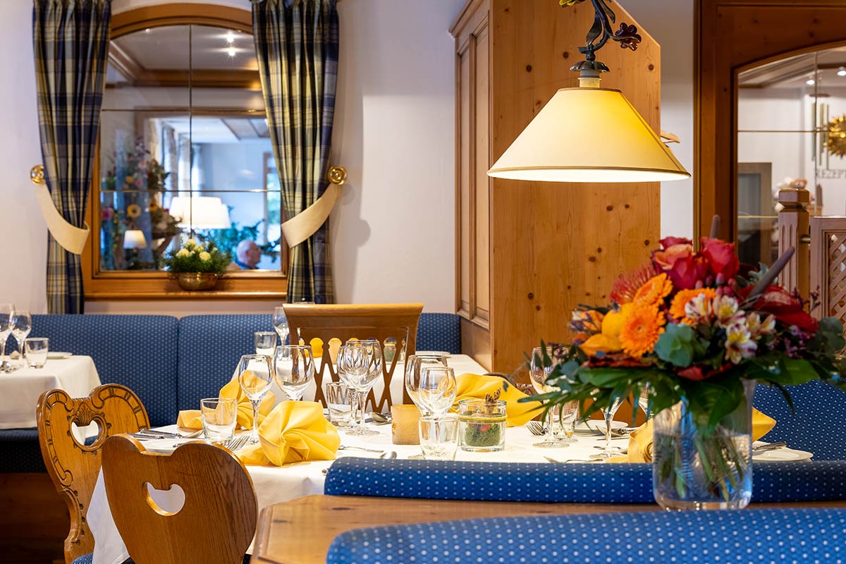 Nahaufnahme eines gedeckten Tisches mit Weingläsern, Blumen und hellblauer gepolsterter Bank unter Holzdecke.