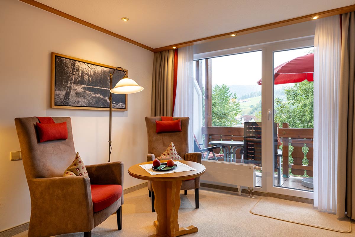 Tisch mit zwei Sesseln vor breiter Fensterfront, Terrasse mit Sonnenschirm im Hintergrund sichtbar.