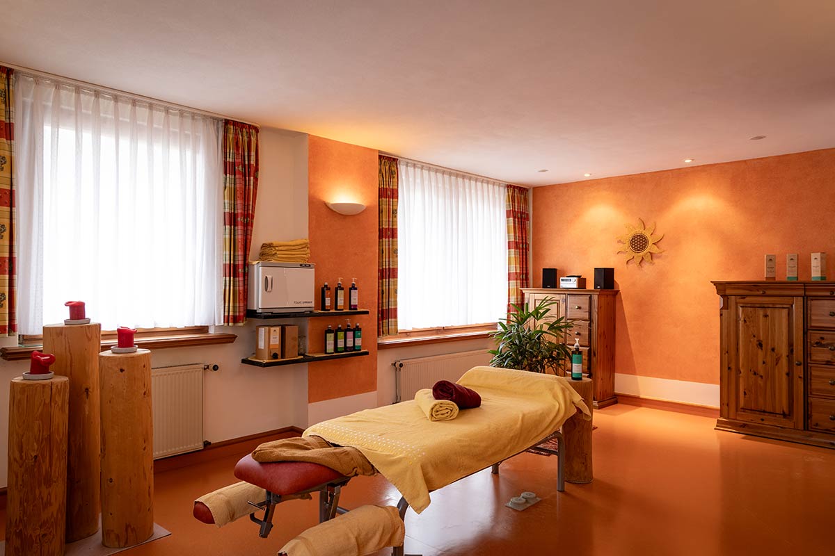 Behandlungsraum mit zwei Massageliegen, Handtücher aufgerollt, heller Boden und Fenster mit Vorhängen.