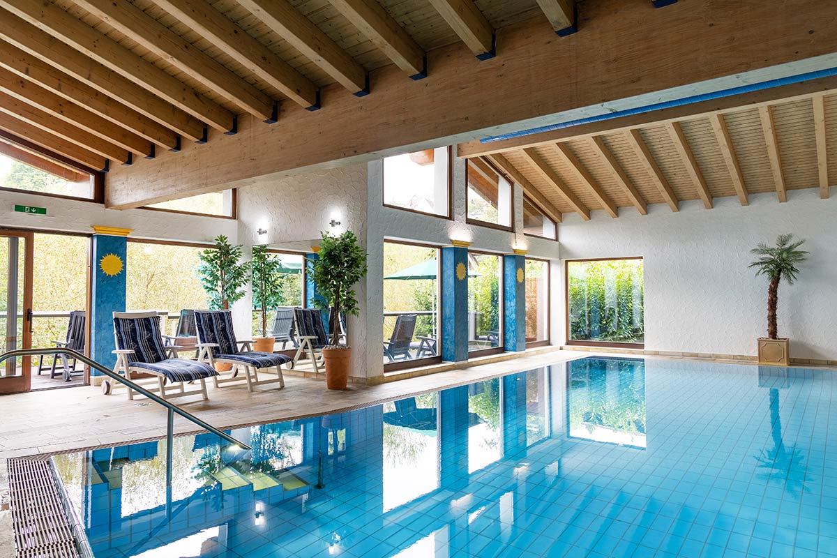 Beheizter Innenpool mit Liegestühlen, Bodenfliesen, durchgehender Fensterfront und offener Dachstruktur.