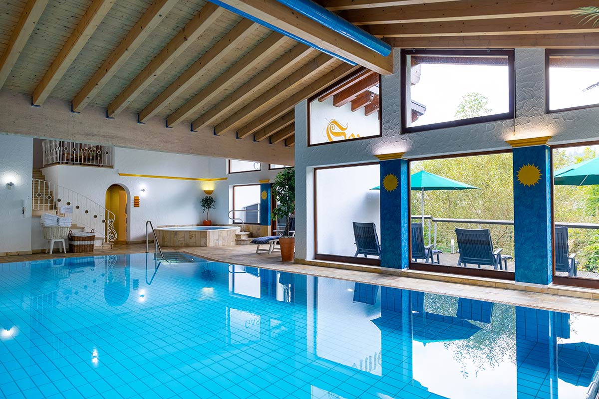Beheizter Innenpool mit Liegestühlen, Bodenfliesen, durchgehender Fensterfront und offener Dachstruktur.