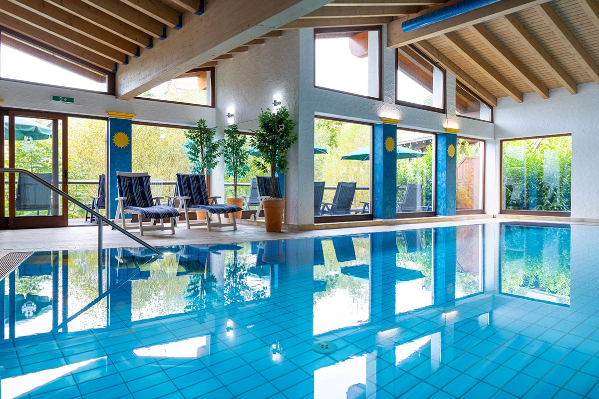 Beheizter Innenpool mit Liegestühlen, Bodenfliesen, durchgehender Fensterfront und offener Dachstruktur.