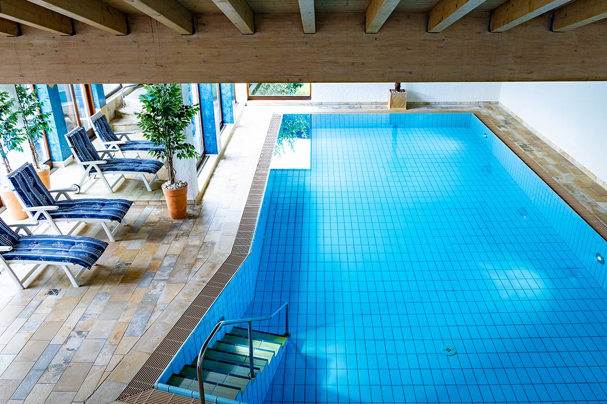 Innenpool mit türkisfarbenen Fliesen, breiten Fenstern auf zwei Seiten, Holzdecke mit dunklen Sichtbalken.