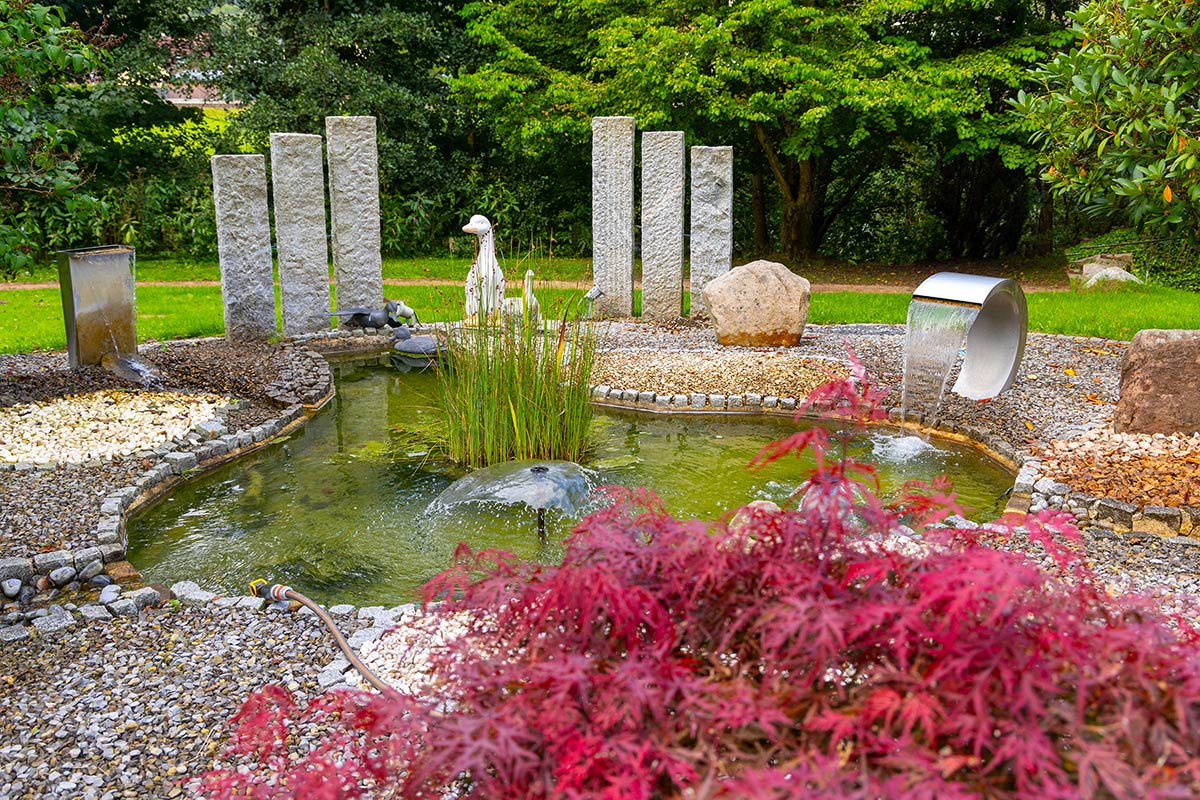 Gartenanlage mit Zierteich, Steinstehlen, Wasserlauf, roten Ahornbüschen und grüner Bepflanzung.