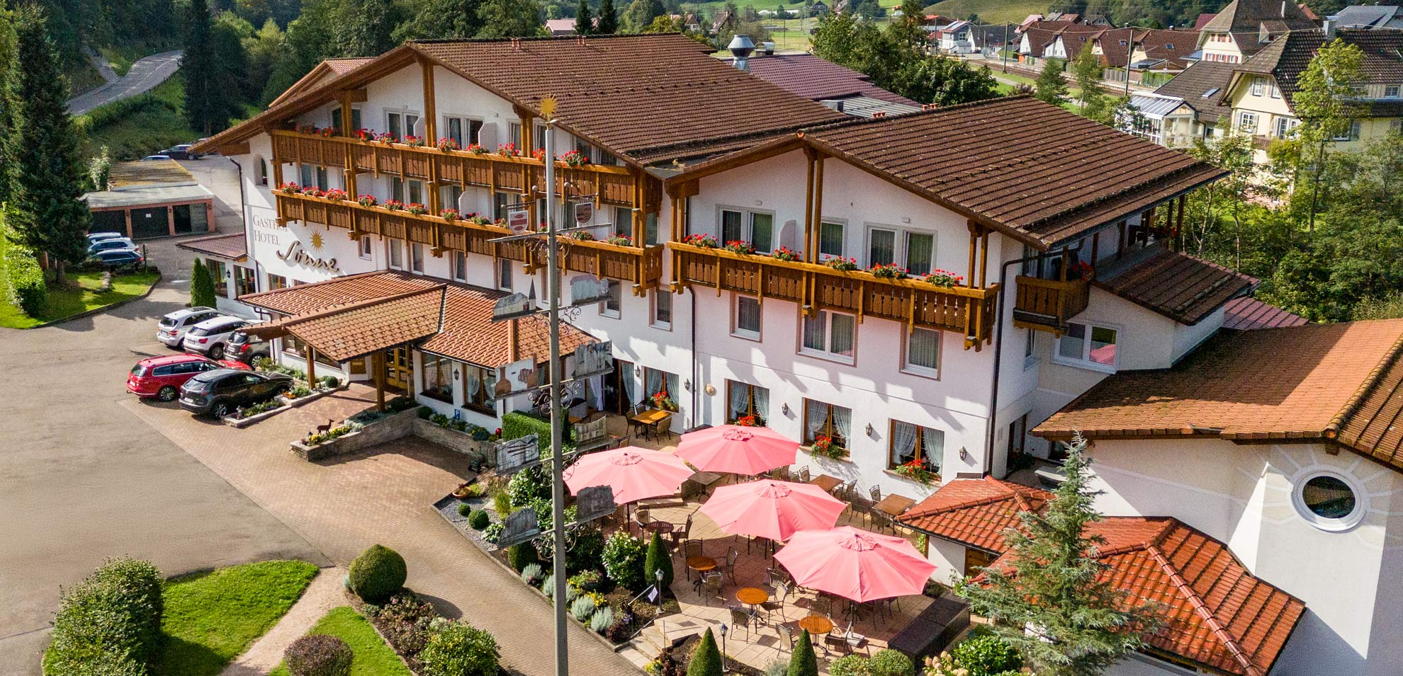 Luftaufnahme des Hotel Sonne Baiersbronn mit mehreren Gebäuden, roten Dächern und rosa Sonnenschirmen auf der Außenterrasse.