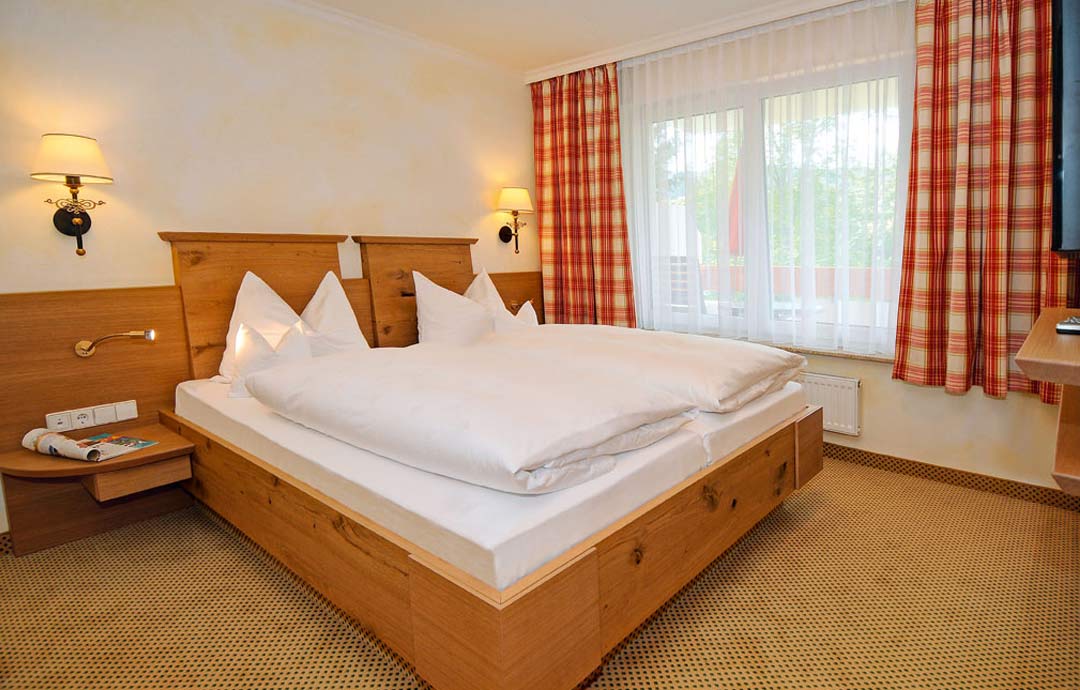 Doppelzimmer Comfort mit großem Doppelbett, weißer Bettwäsche, zwei Nachttischen mit Lampen, Holzboden und Fenster.
