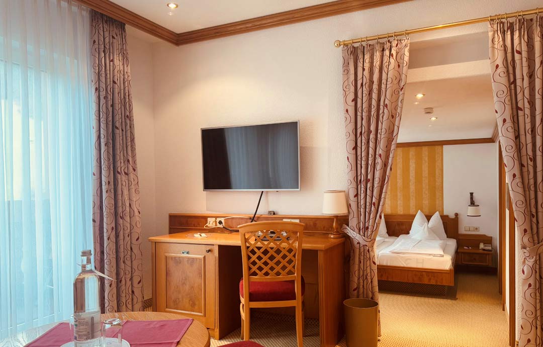 Hotelzimmer Deluxe im Hotel Sonne Baiersbronn mit Schreibtisch, zwei Stühlen, Flachbildfernseher und bodenlangem Vorhang.