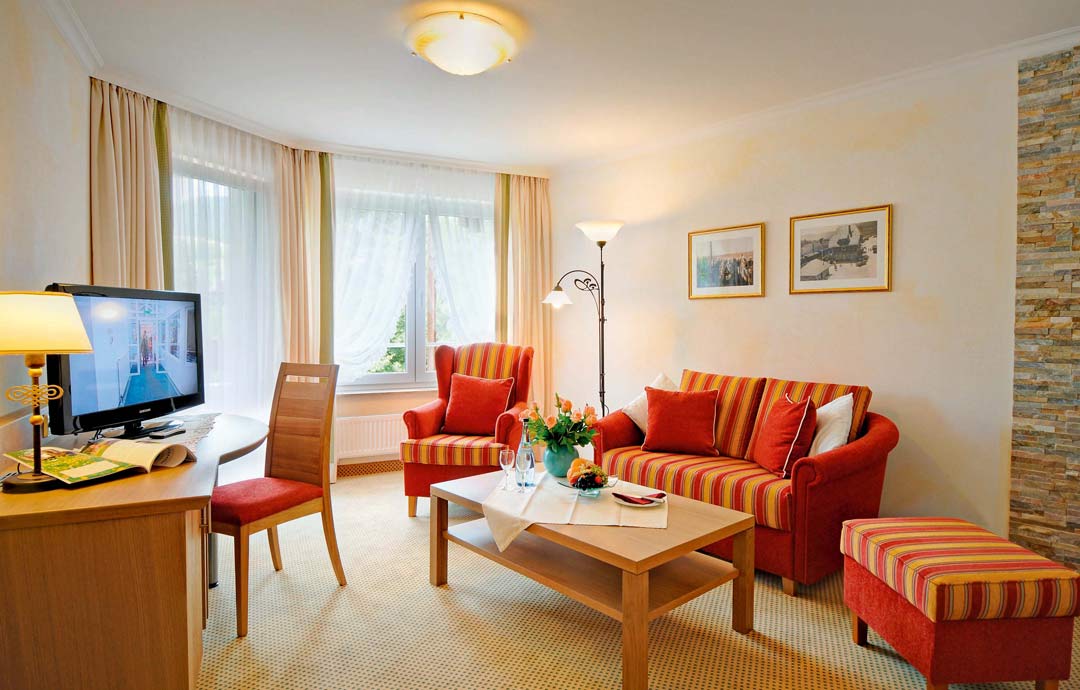 Wohnbereich Suite mit gemustertem Sofa, Sessel, Couchtisch, Vorhängen und Bildern an der Wand, Tageslicht von links.