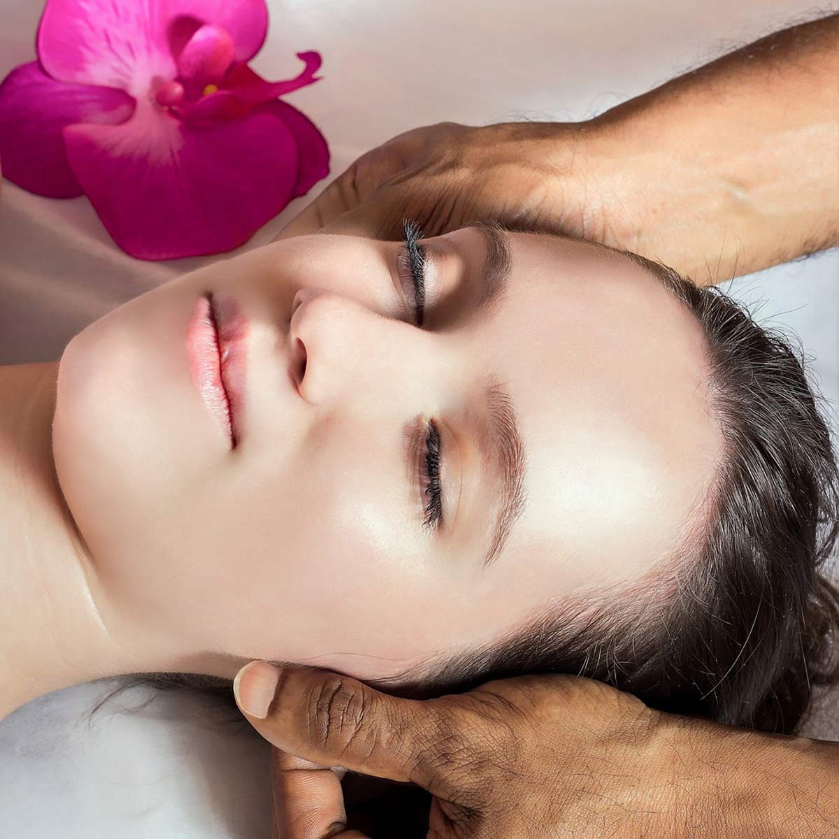 Frau liegt auf einer Spa-Liege, Augen geschlossen, erhält ruhige Gesichtsmassage mit beiden Händen, neutrale Lichtstimmung.