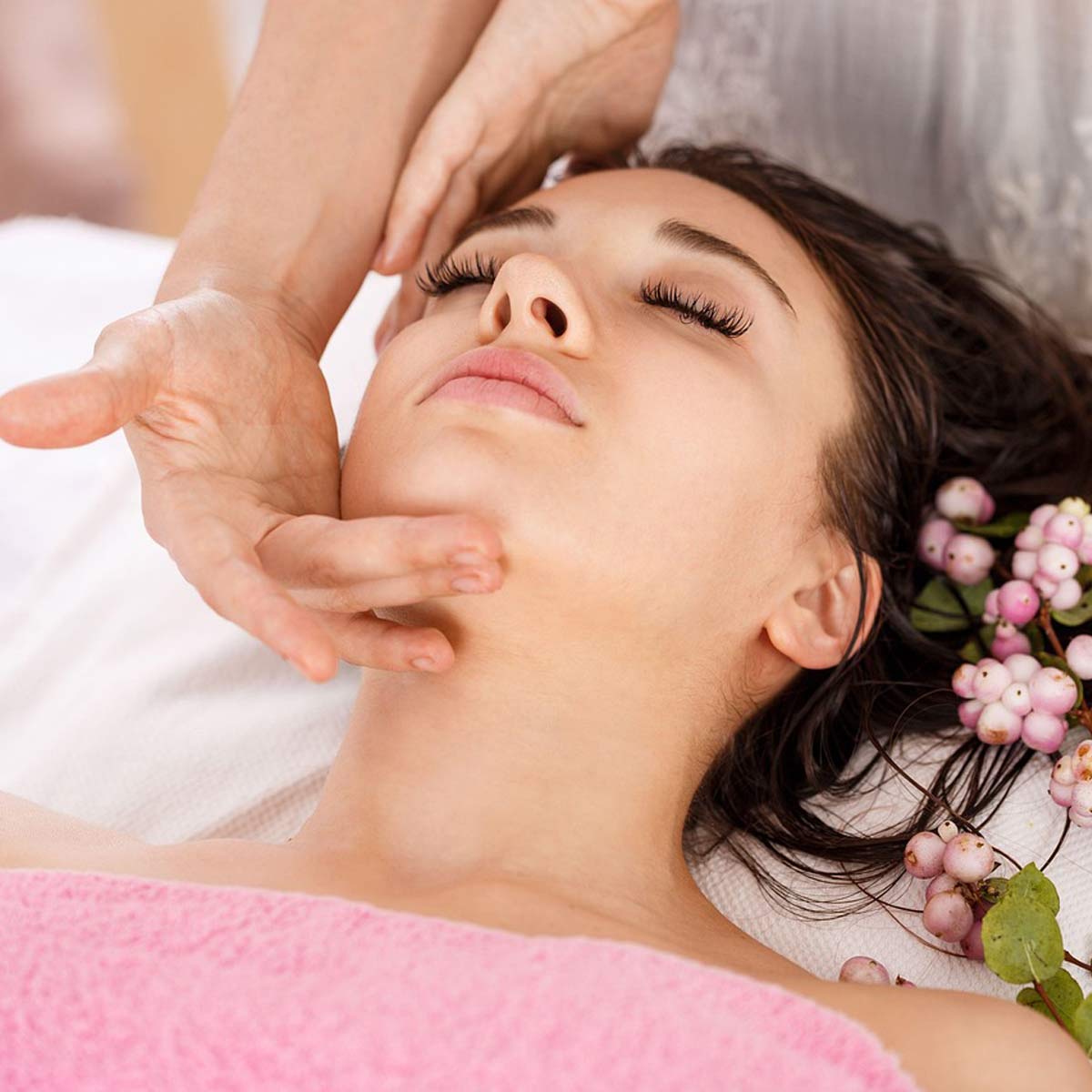 Gesichtsmassage mit leichtem Druck entlang der Kieferpartie, Frau liegt unter rosa Handtuch mit Blüten.