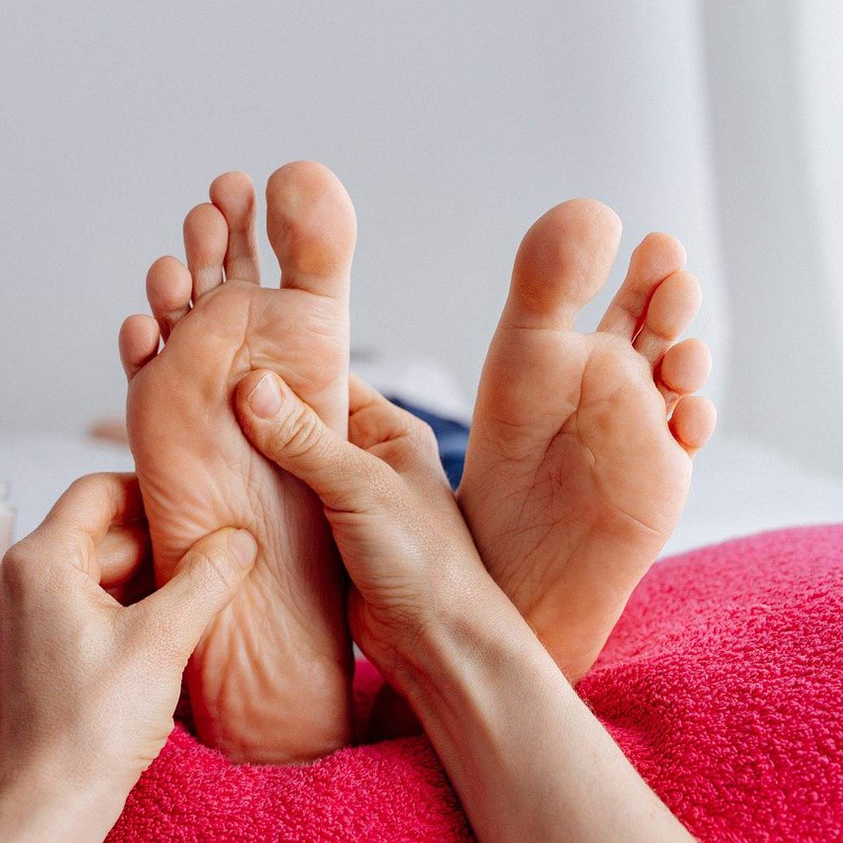 Fußmassage mit zwei Händen, eine Person liegt auf rosa Unterlage, weiches Licht, Nähe zu Zehen erkennbar.
