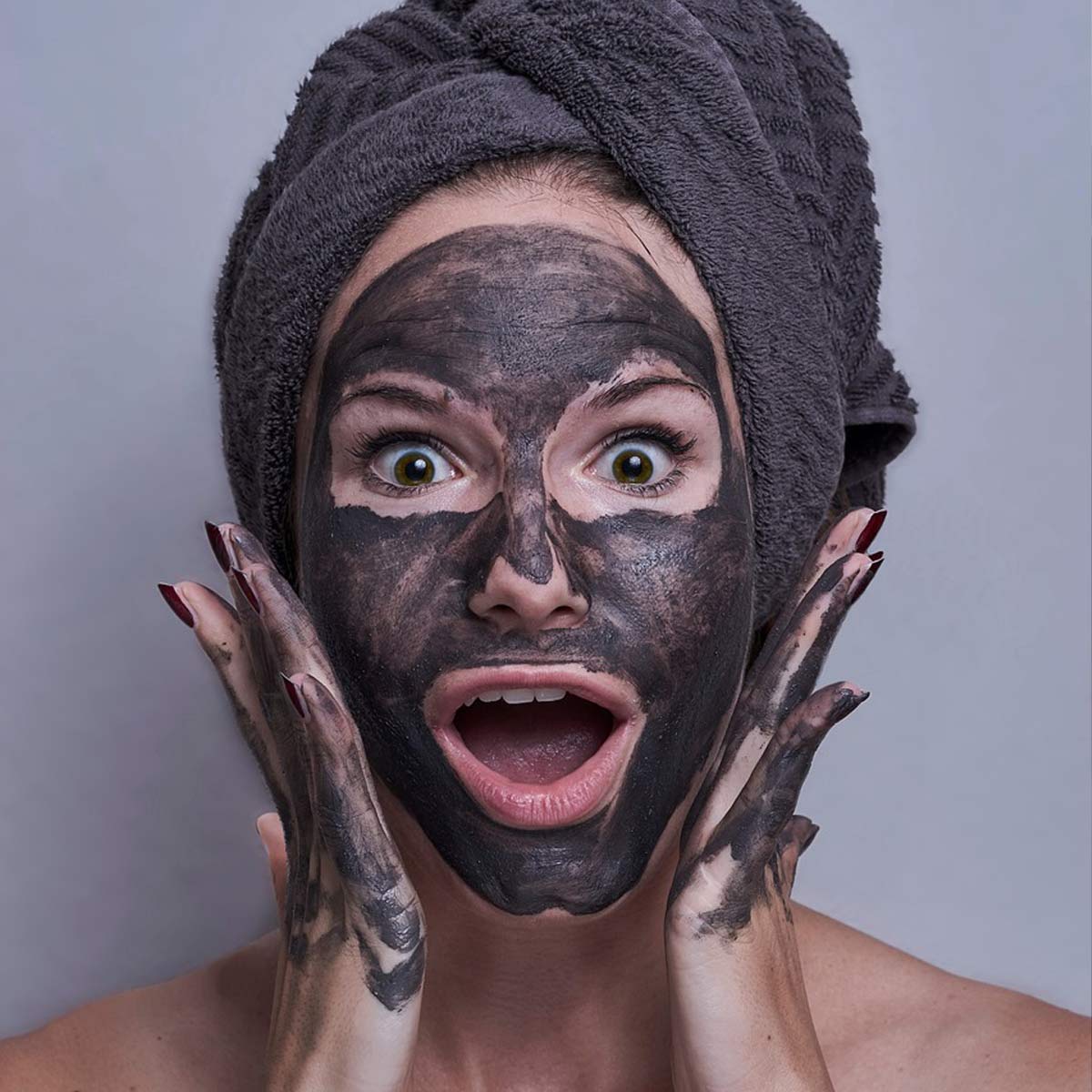 Frau mit dunkler Gesichtsmaske, Handtuchturban und weit aufgerissenen Augen hält lachend beide Hände an ihr Gesicht.