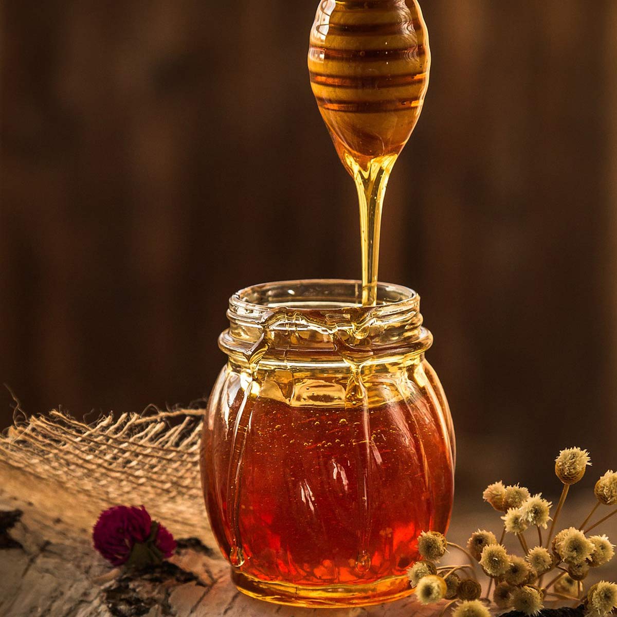 Honig läuft aus Holzlöffel in ein Glas, daneben Zimtstange, Trockenblüten und rustikale Leinentischdecke.