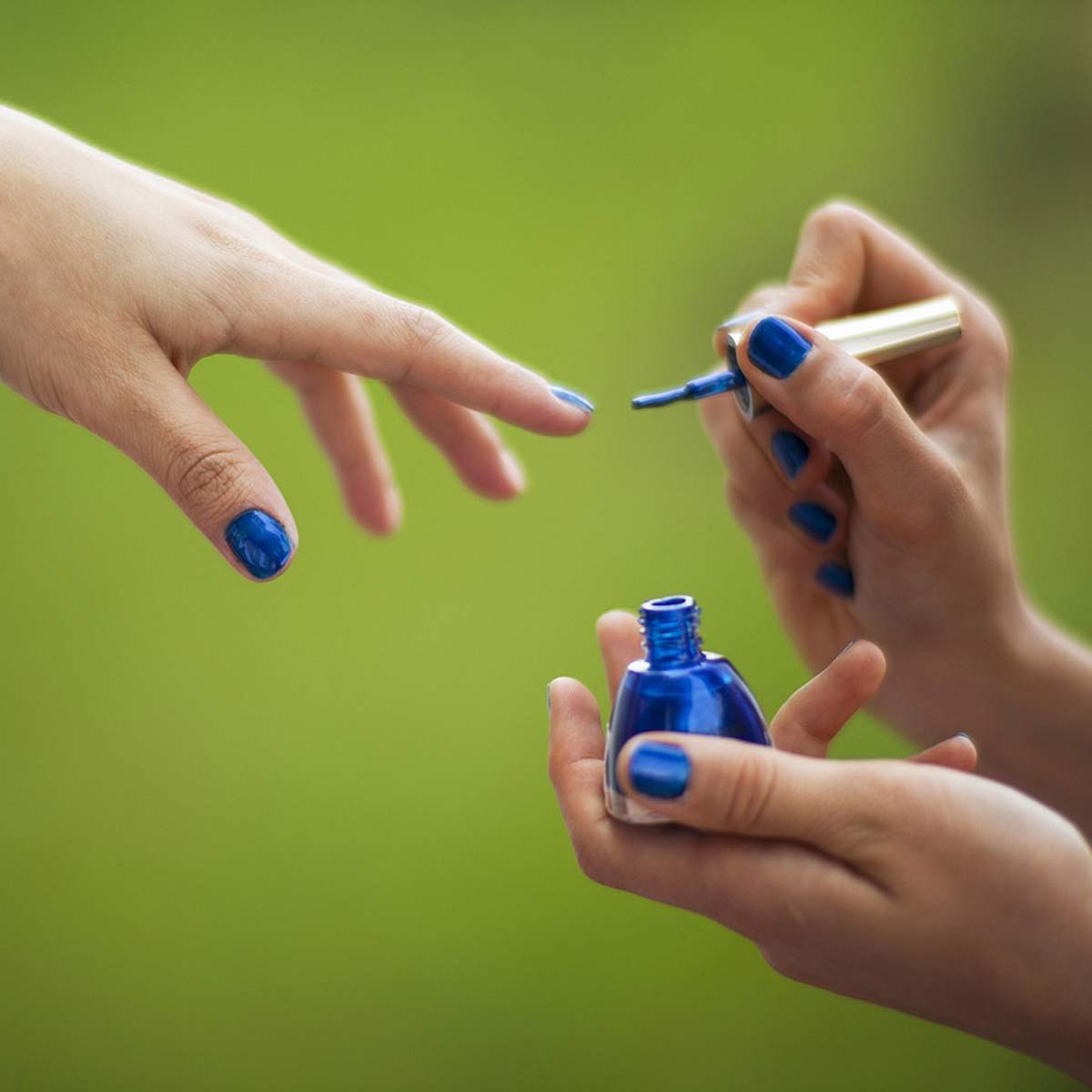 Nahaufnahme einer Hand, die mit einem Pinsel blauen Nagellack aus einer Glasflasche tropft; Hintergrund unscharf grün.