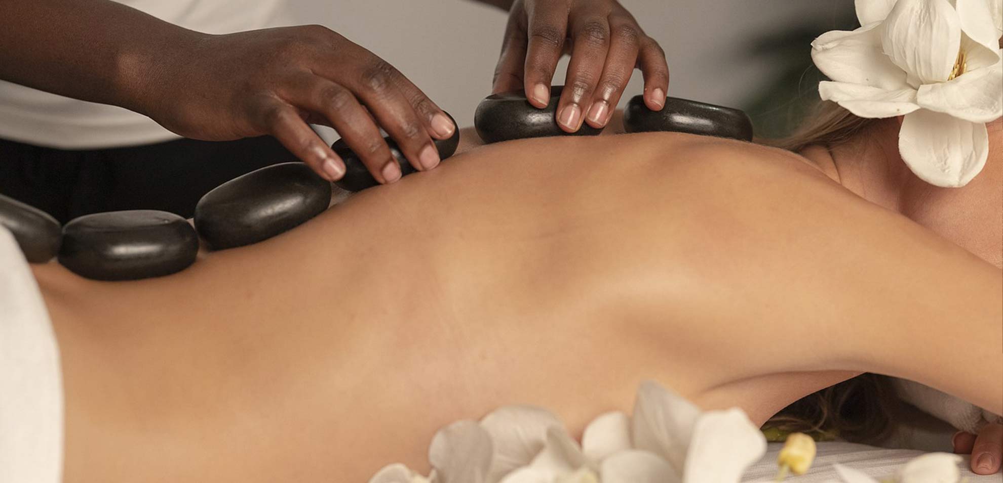 Hot Stone Massage mit warmem Öl, Hände streichen sanft über die Haut, liegende Person auf weißer Liege mit Blüten.